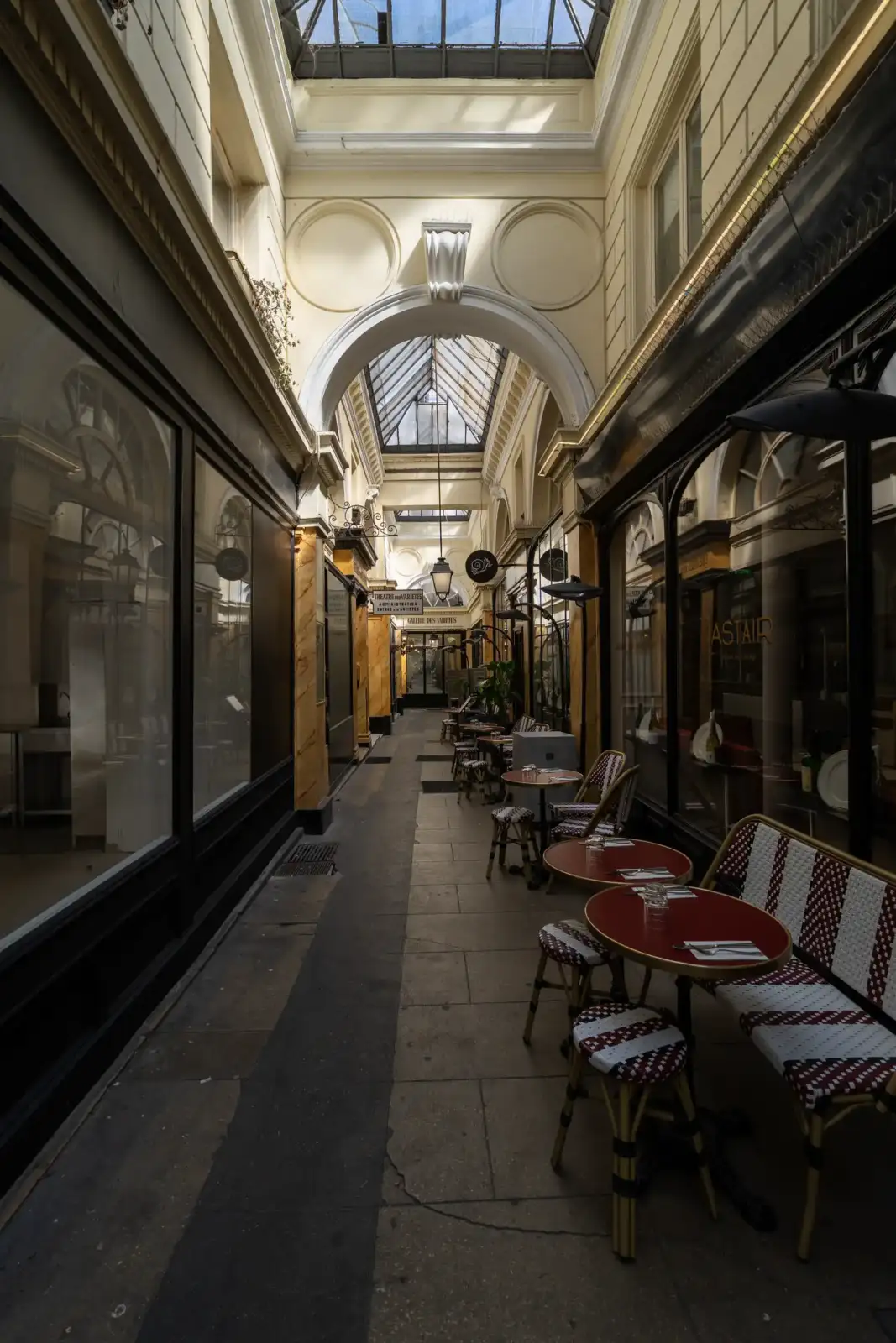 Vue intérieure du passage des Panoramas illuminé, bordé de vitrines et de petites tables de café.