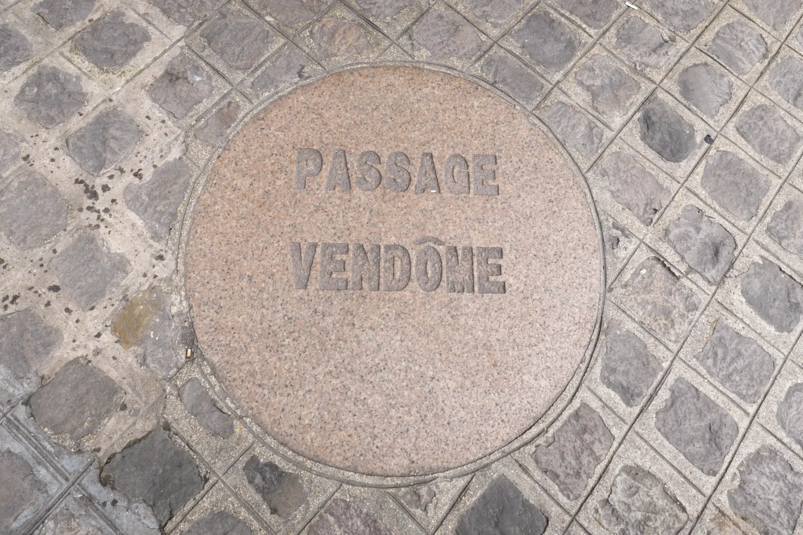 Une plaque circulaire en pierre indiquant Passage Vendôme insérée dans un sol pavé parisien.