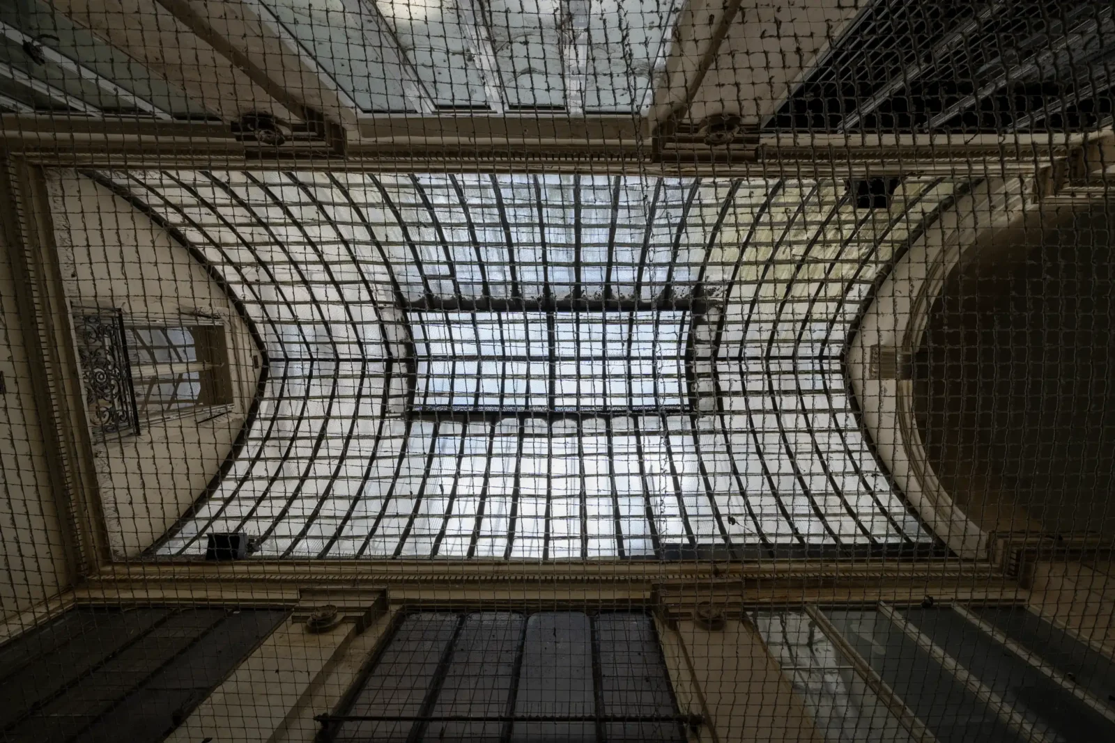 Vue en contre-plongée de la verrière ancienne du Passage Vendôme protégée par un filet.