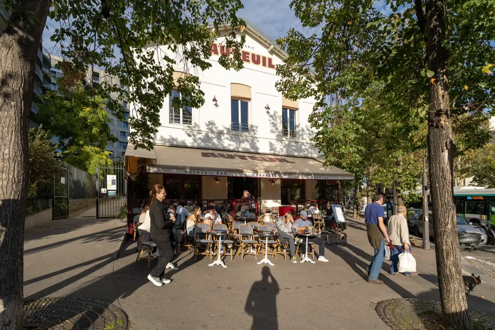 Une brasserie parisienne lumineuse avec une terrasse animée et des clients attablés sous les arbres.