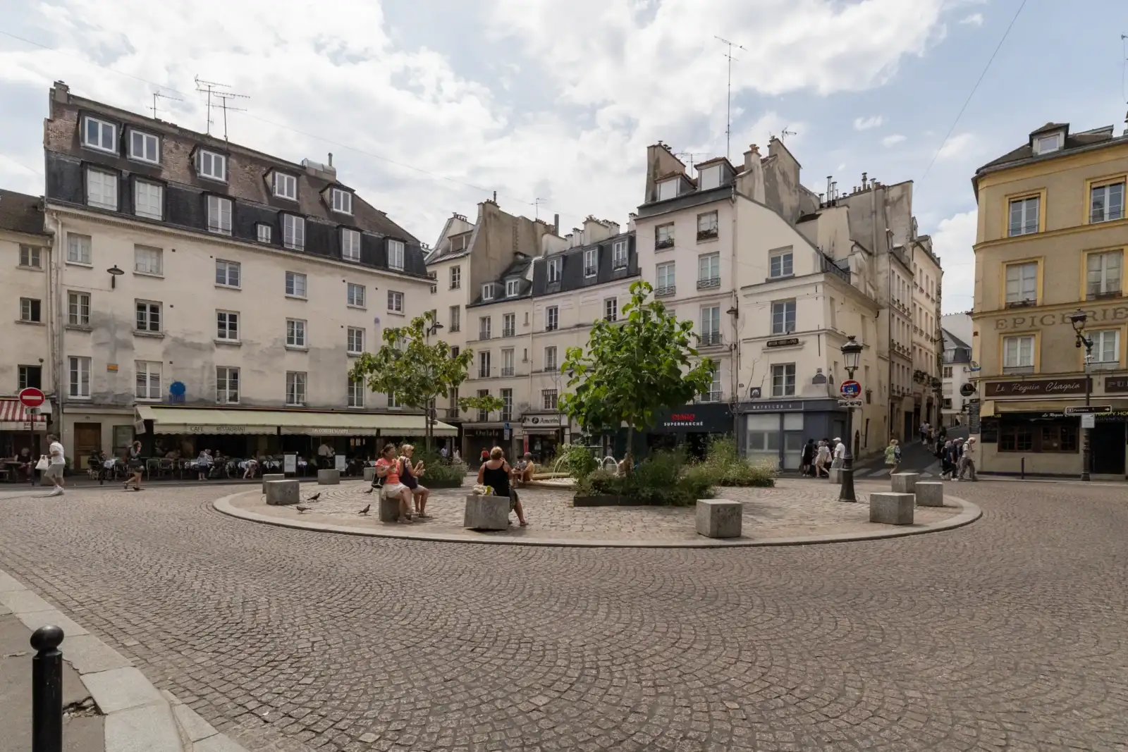 Une place pavée entourée d'immeubles parisiens anciens avec des terrasses de cafés animées.