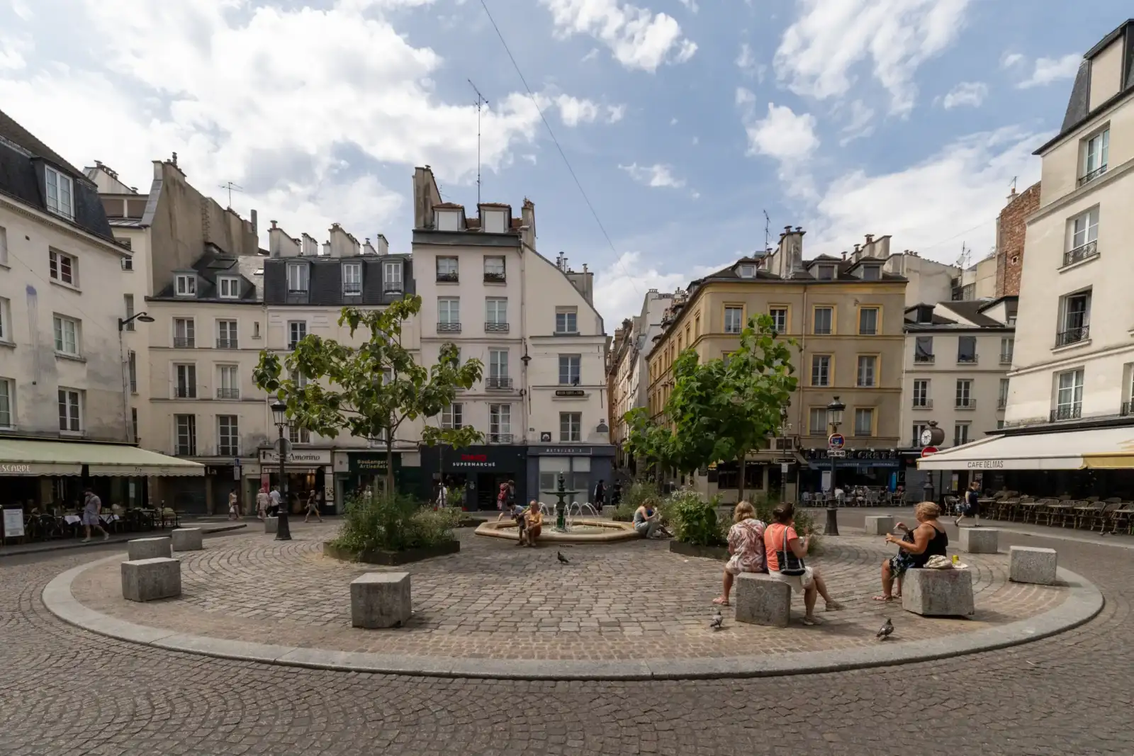 Une place pavée animée à Paris avec des immeubles anciens et des arbres.