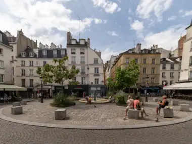 Une place pavée animée à Paris avec des immeubles anciens et des arbres.