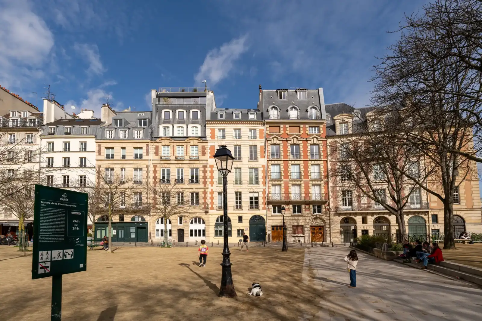 Une place parisienne ensoleillée bordée d'immeubles classiques avec des gens et un chien.