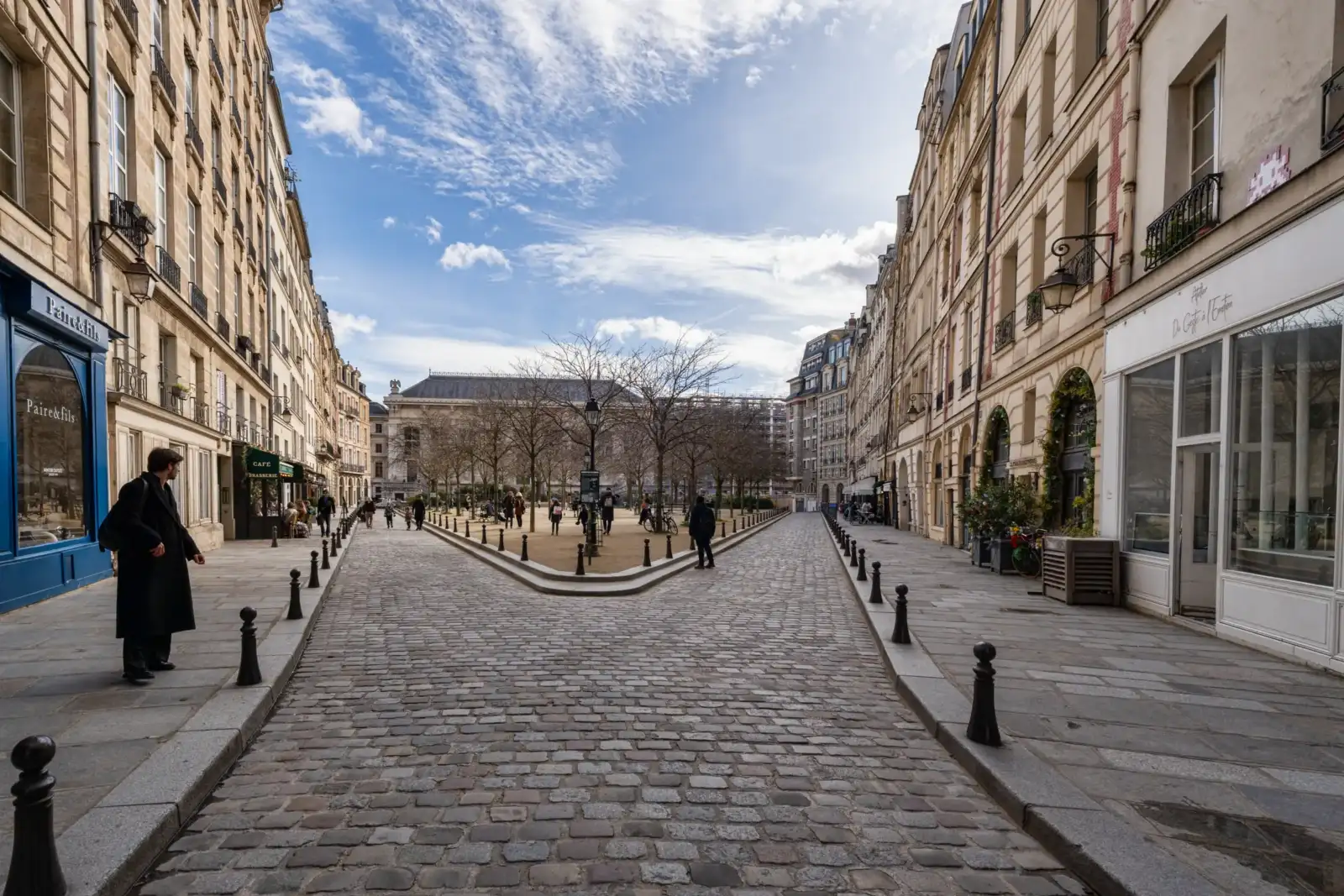 Une rue pavée mène vers la Place Dauphine bordée d'immeubles parisiens sous un ciel bleu.