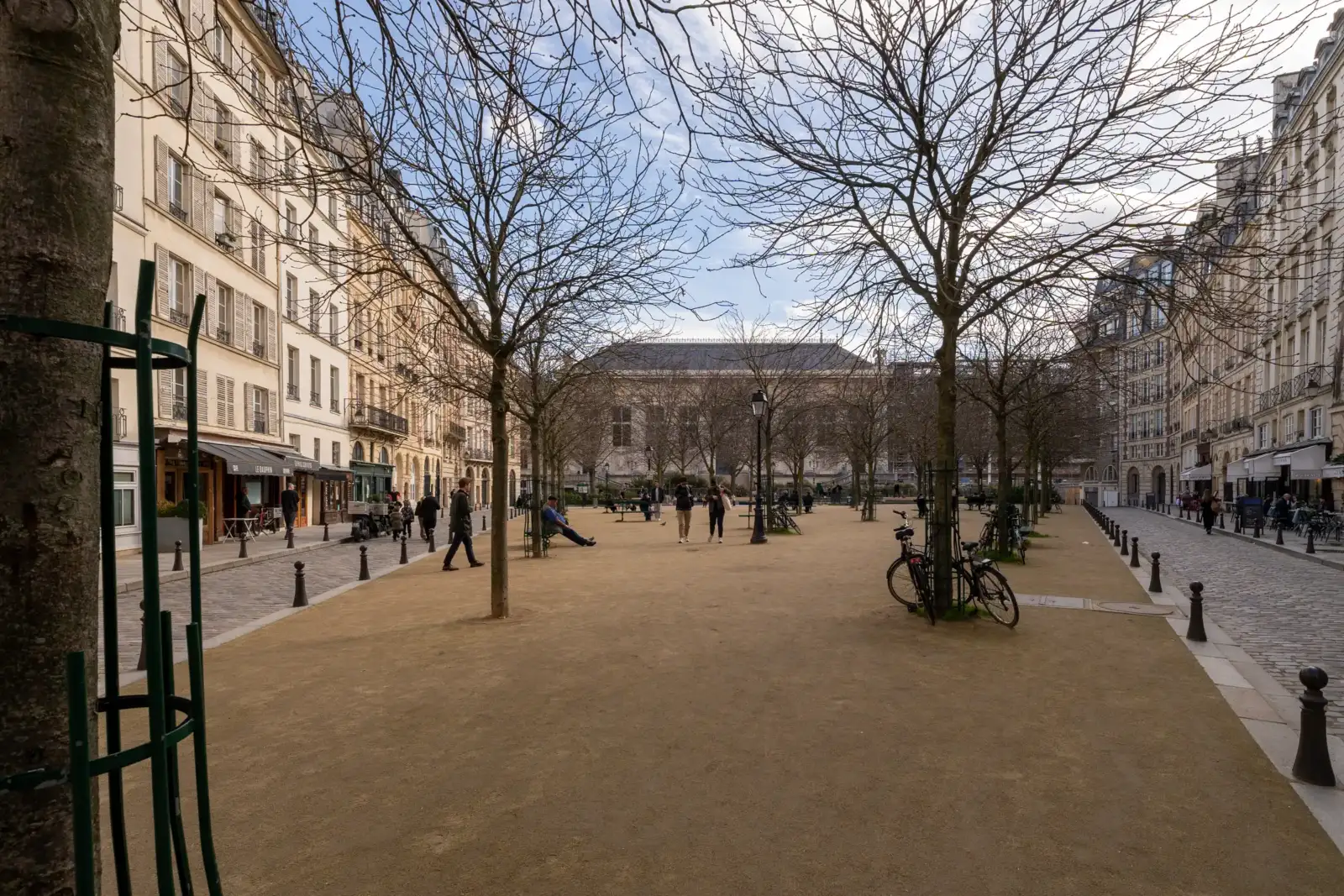Vue sur la place Dauphine à Paris avec ses arbres dénudés et passants.