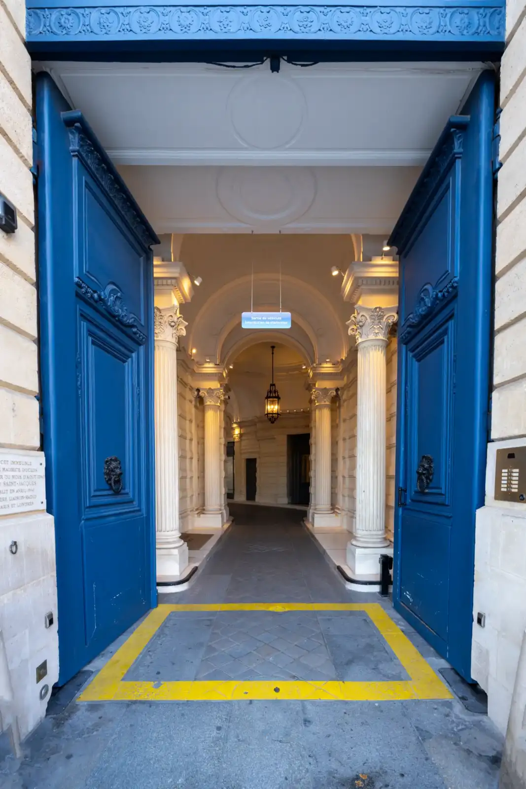 Grandes portes bleues ouvertes sur un élégant passage voûté bordé de colonnes classiques parisiennes.