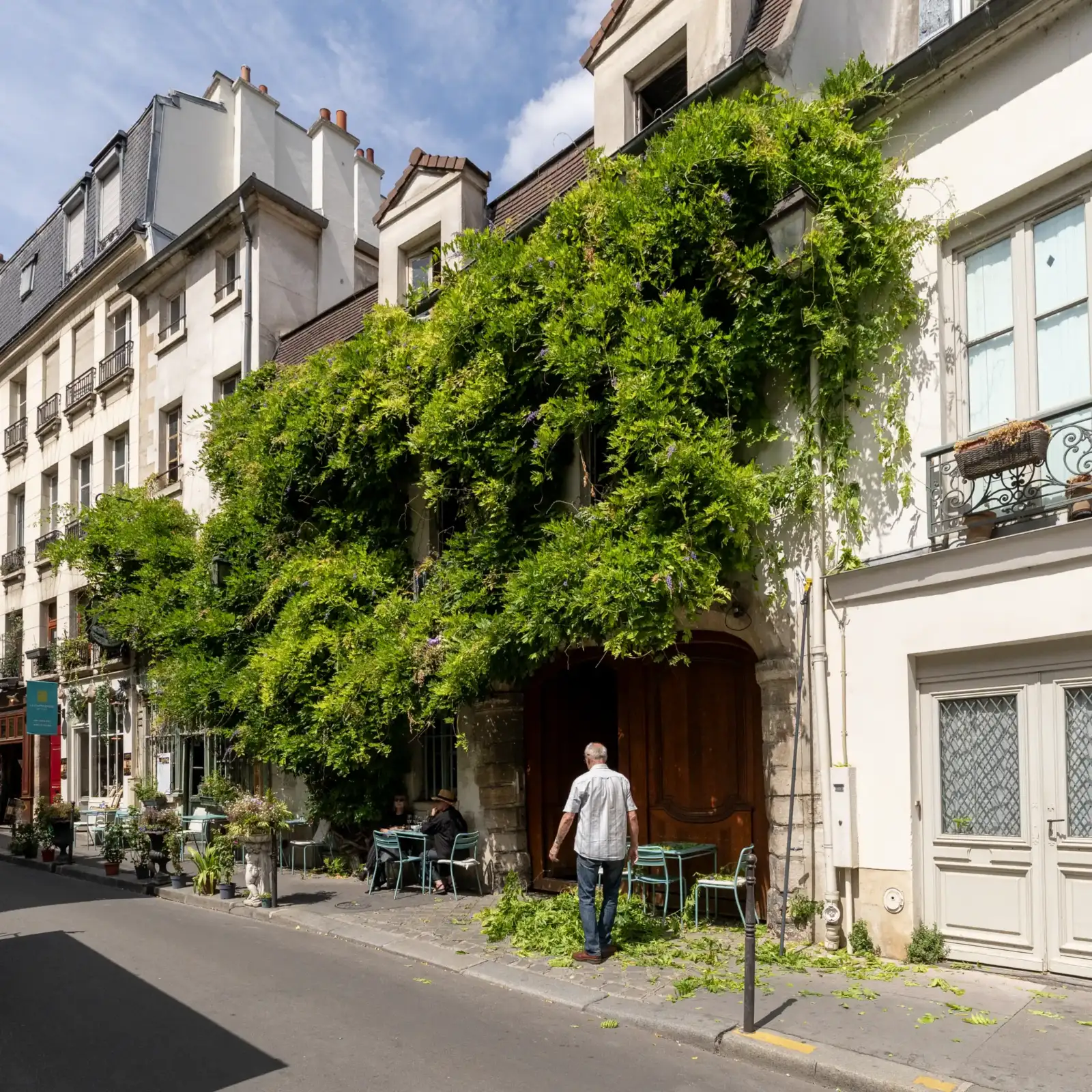 Un homme balaie des feuilles vertes sur le trottoir devant une façade parisienne luxuriante.