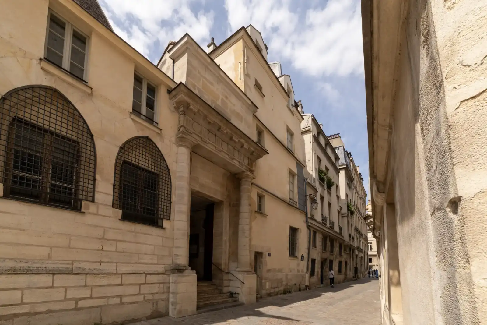 Une ruelle parisienne étroite bordée de façades historiques en pierre claire sous un ciel nuageux.
