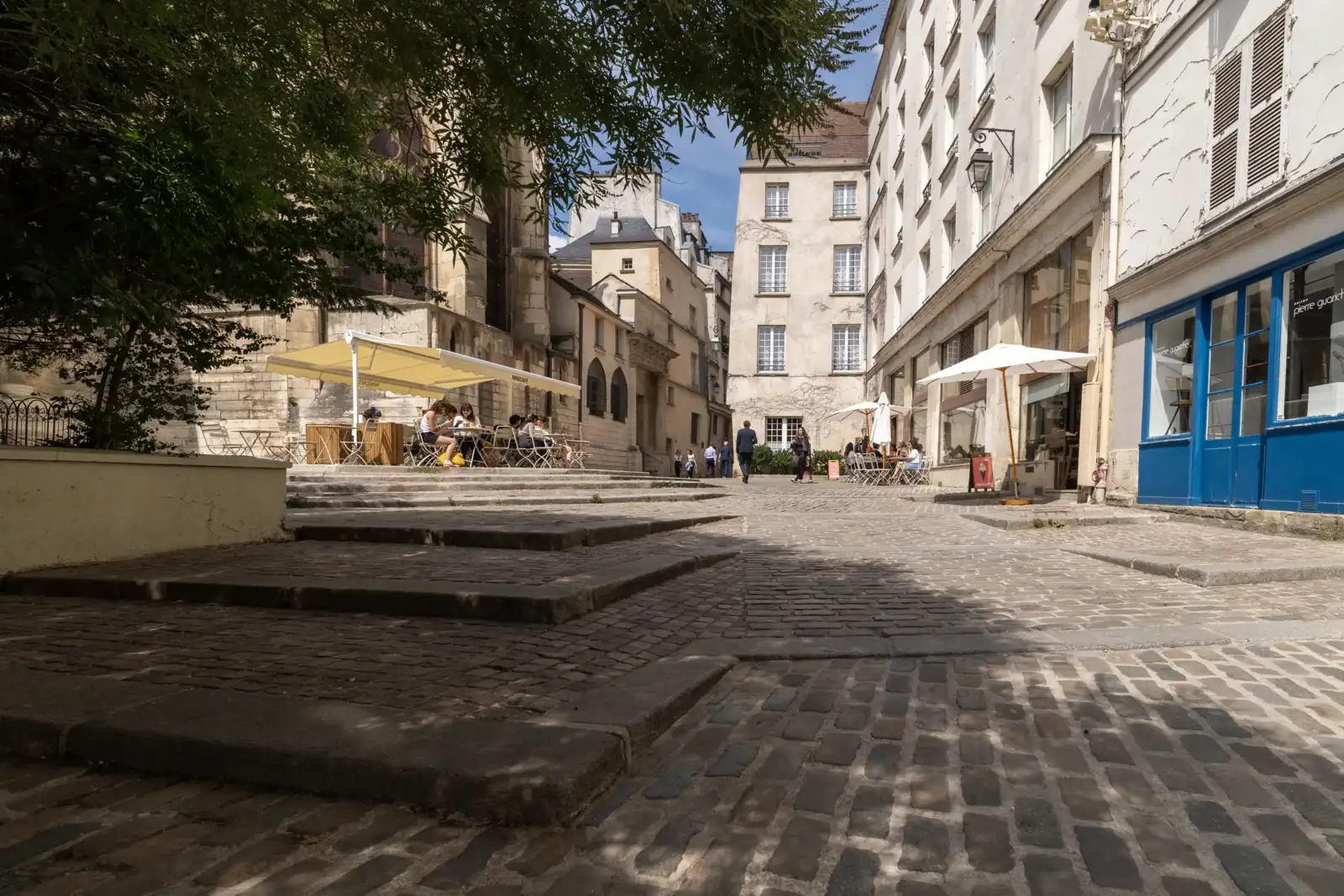 Une rue pavée pittoresque à Paris avec des terrasses de café ensoleillées et des bâtiments historiques.