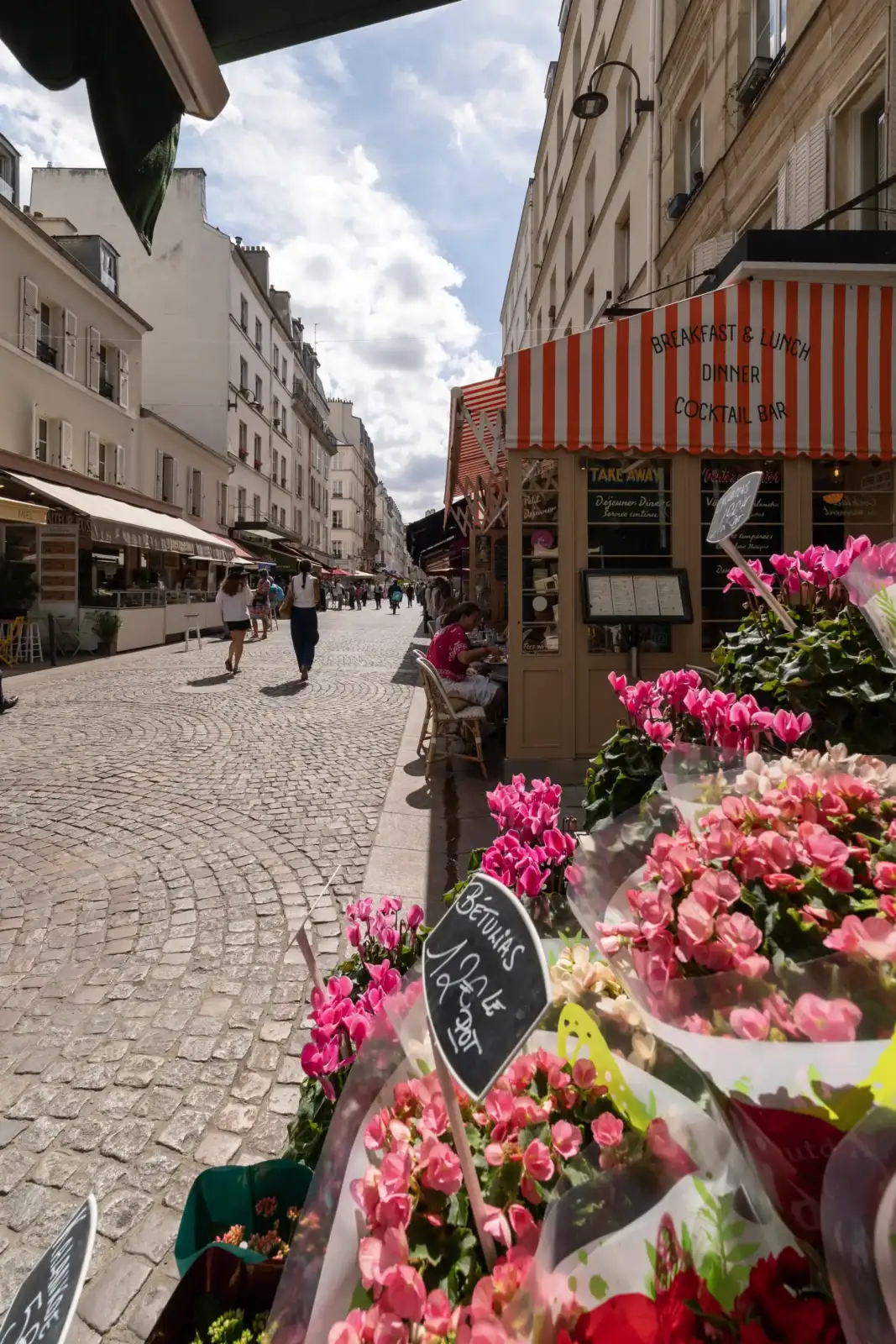 Une rue pavée animée à Paris avec des terrasses de café et des fleurs.