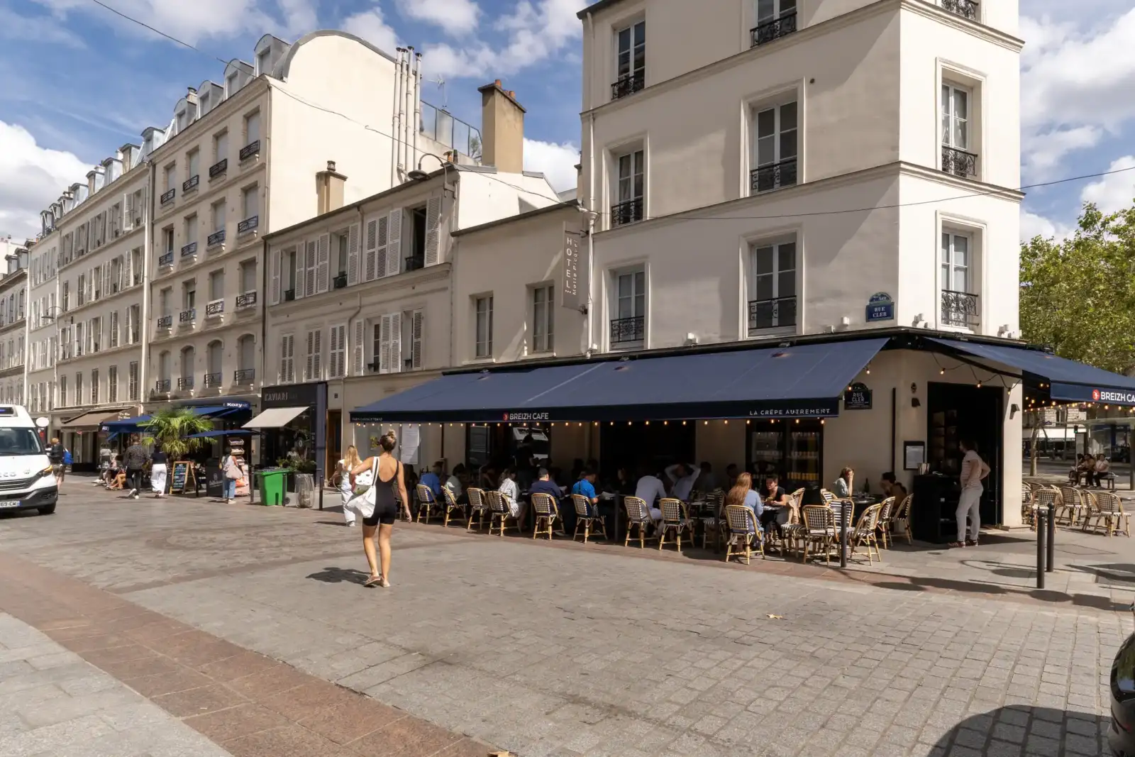 Une femme marche sur une rue piétonne pavée bordée d'immeubles parisiens et de cafés.