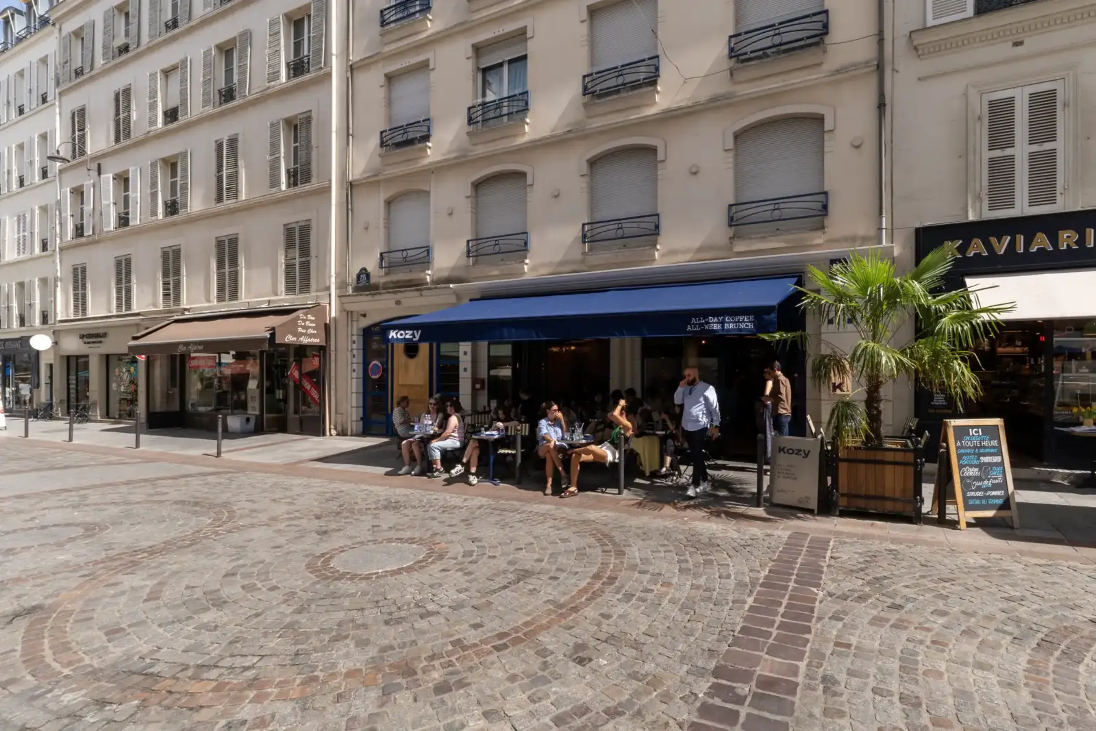 Façade de immeubles parisiens anciens avec des commerces et un café animé en terrasse.