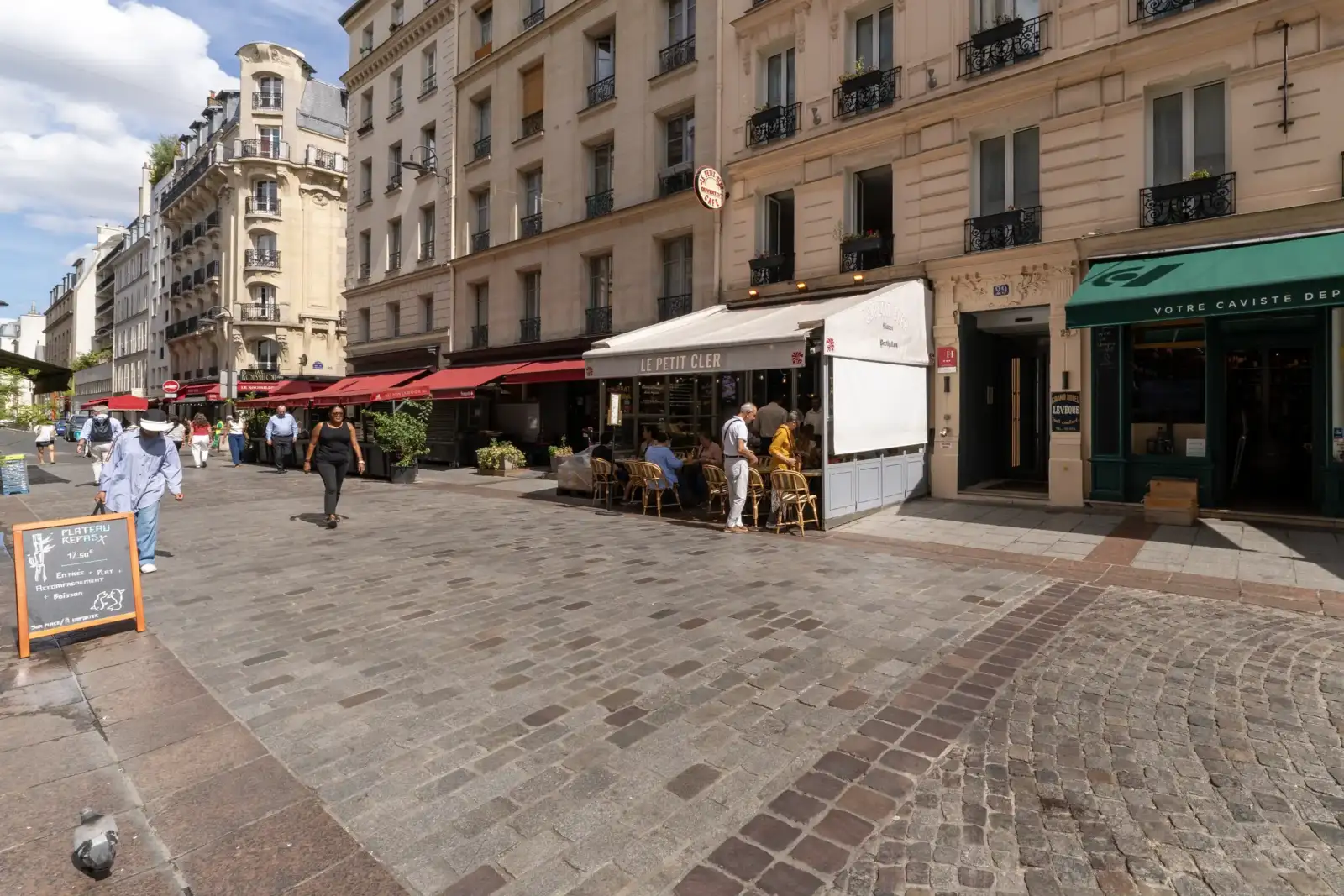 Une rue pavée animée de Paris bordée de bâtiments haussmanniens et de terrasses.