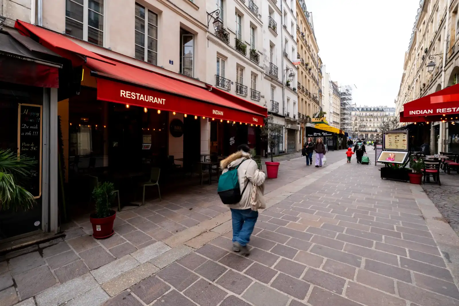 Une rue pavée de Paris avec des restaurants aux stores rouges et des passants.