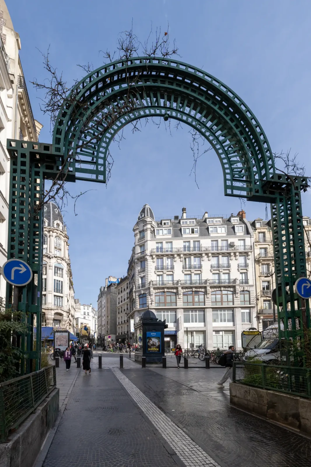 Une arche métallique verte marque l'entrée piétonne d'une rue animée à Paris.