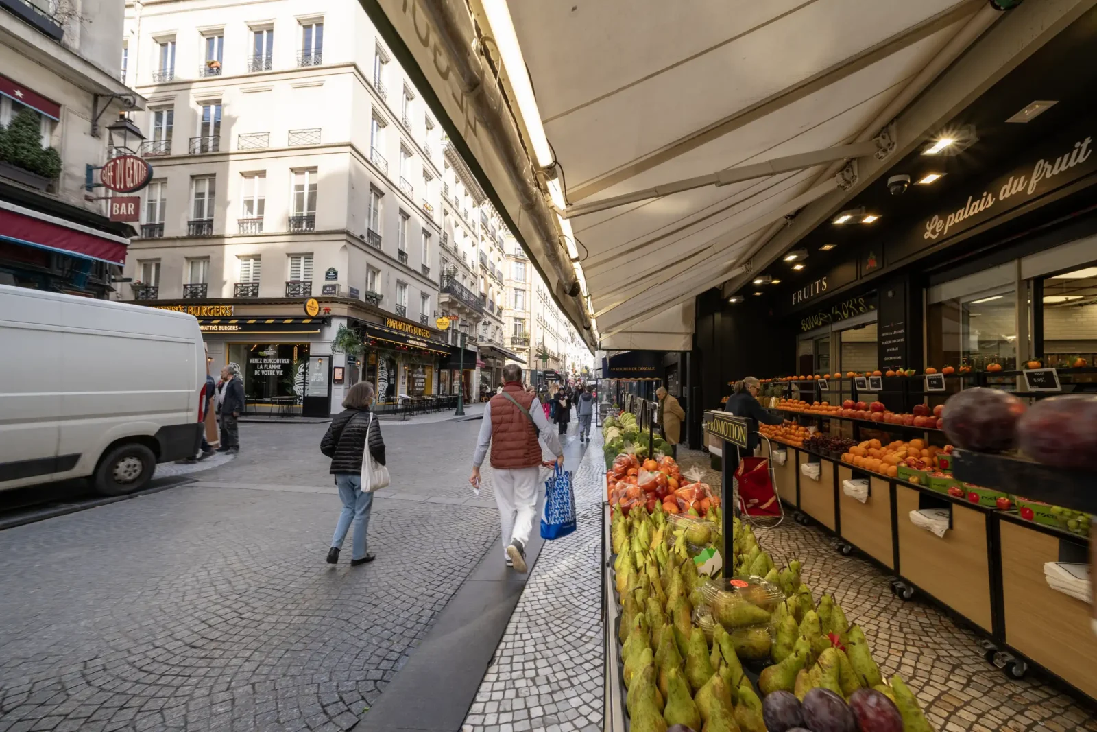 Des passants marchent dans la rue Montorgueil devant un étalage de fruits frais parisiens.