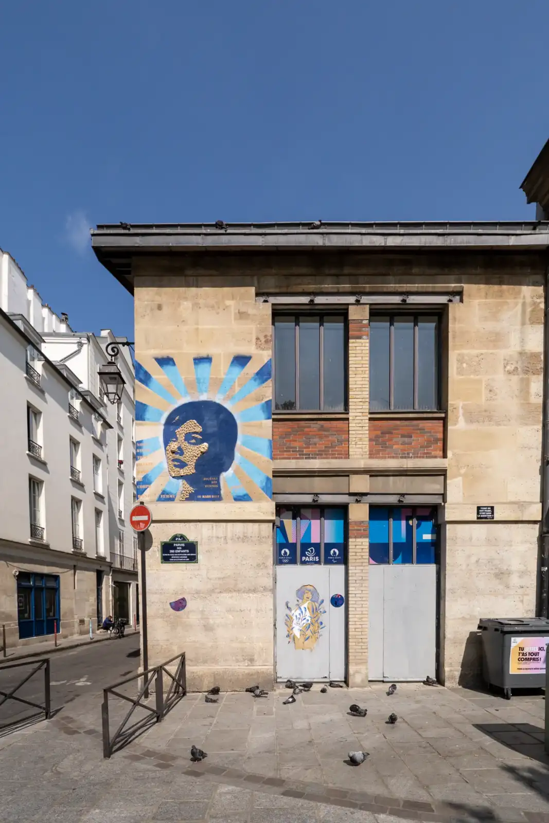 Une fresque murale bleue et dorée représentant un visage féminin orne la façade.