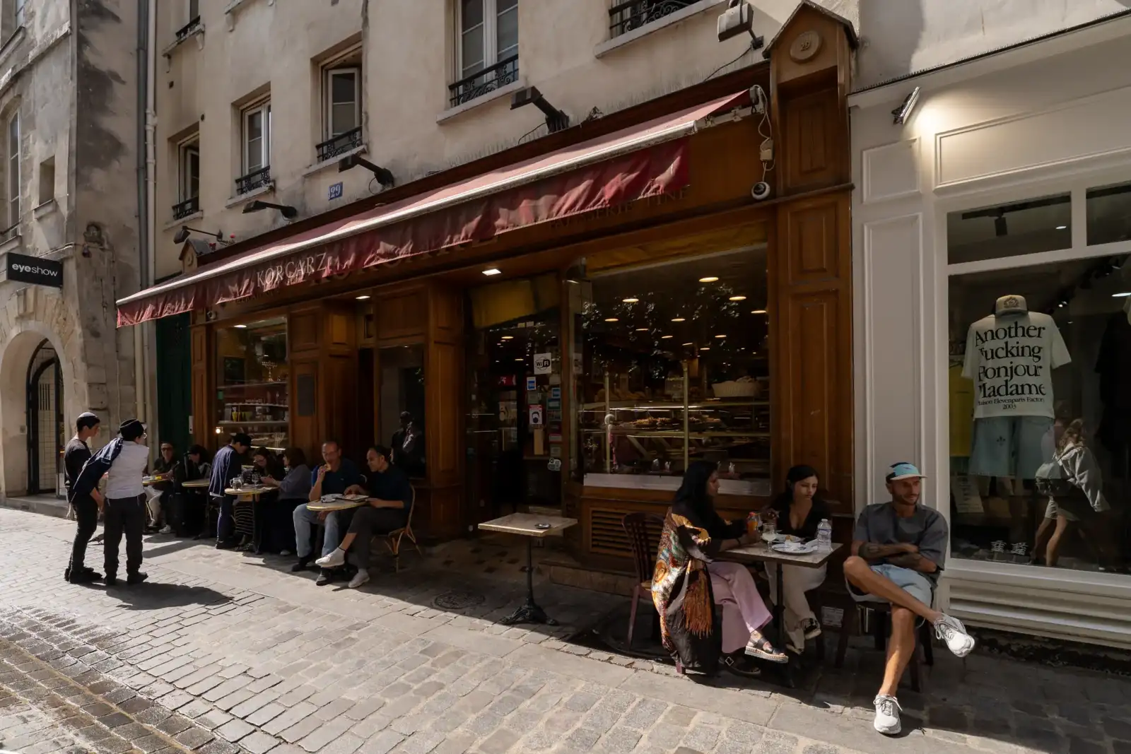Une terrasse animée devant la devanture en bois d'une boulangerie parisienne ensoleillée.