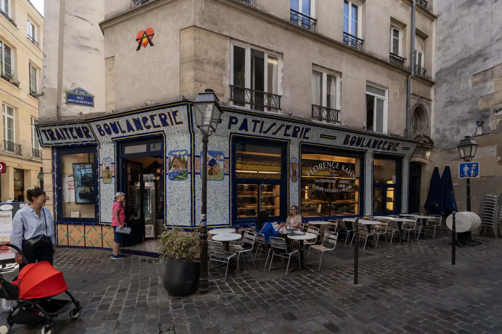 Une boulangerie traditionnelle à la façade carrelée bleue dans une rue pavée parisienne animée.