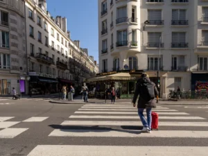 Un voyageur avec une valise rouge traverse une rue ensoleillée du centre de Paris.