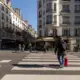 Un voyageur avec une valise rouge traverse une rue ensoleillée du centre de Paris.