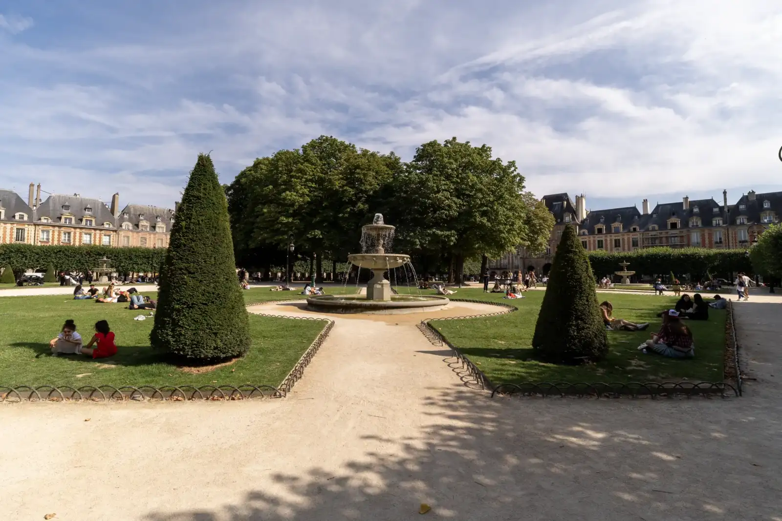 Une fontaine au milieu d’un square parisien arboré avec des pelouses et des passants.