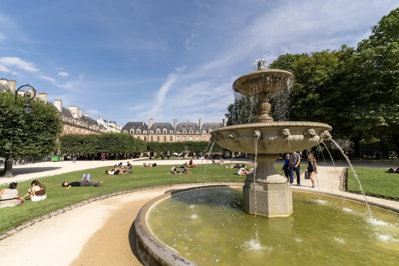 Une fontaine en pierre au premier plan et des personnes profitant de la pelouse ensoleillée.