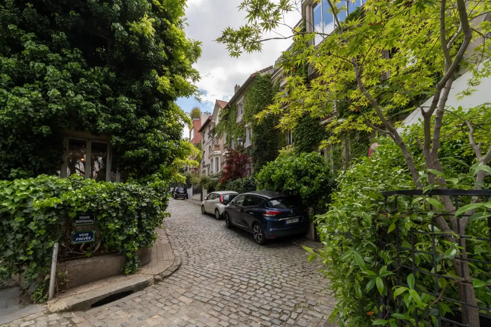 Une rue pavée bordée de maisons verdoyantes et de voitures stationnées au Square Montsouris.