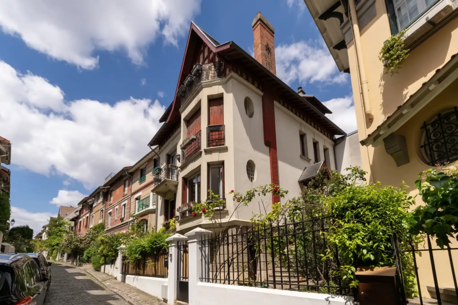 Une ruelle pavée borde une maison ancienne aux volets rouges sous un ciel nuageux.