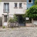 Un vélo violet est garé devant une maison pavée végétalisée.
