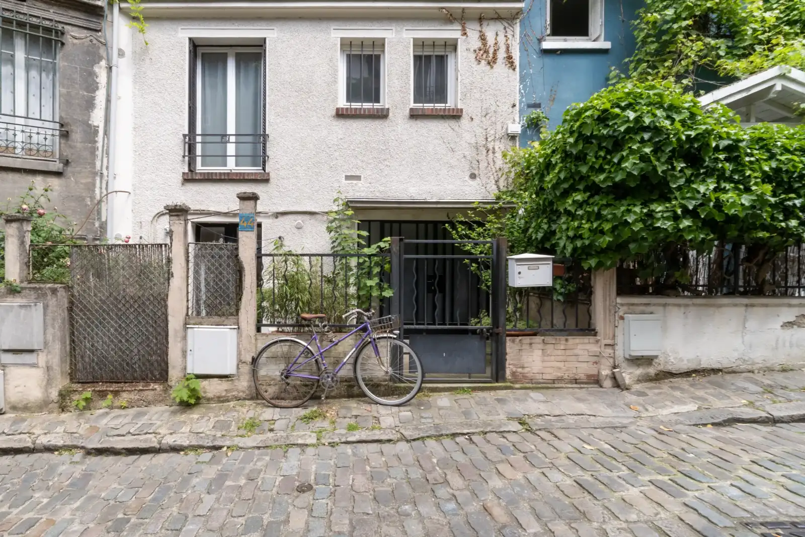 Un vélo violet est garé devant une maison pavée végétalisée.