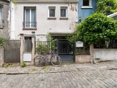 Un vélo violet est garé devant une maison pavée végétalisée.