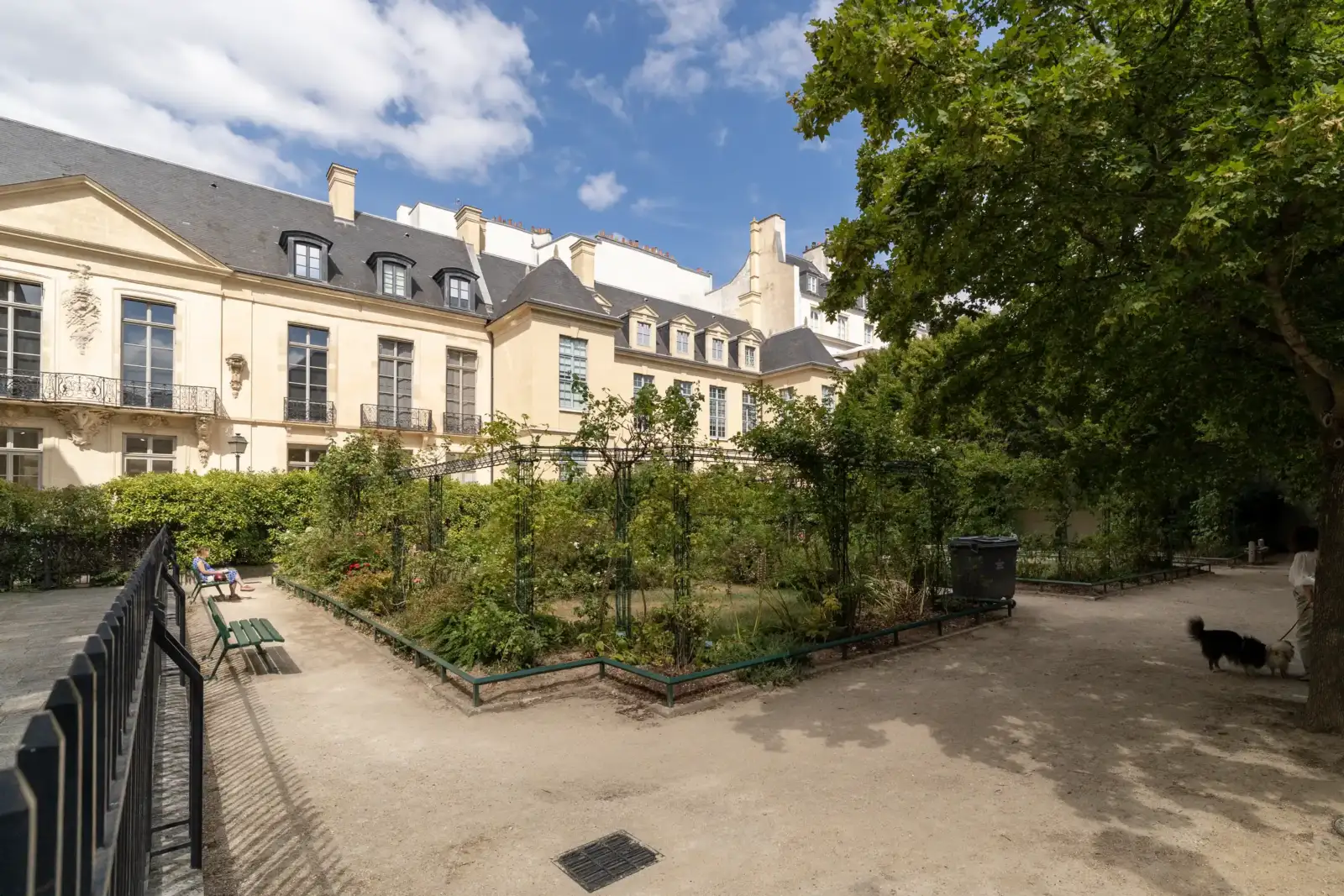Un square parisien verdoyant bordé d'immeubles anciens sous un ciel bleu nuageux.