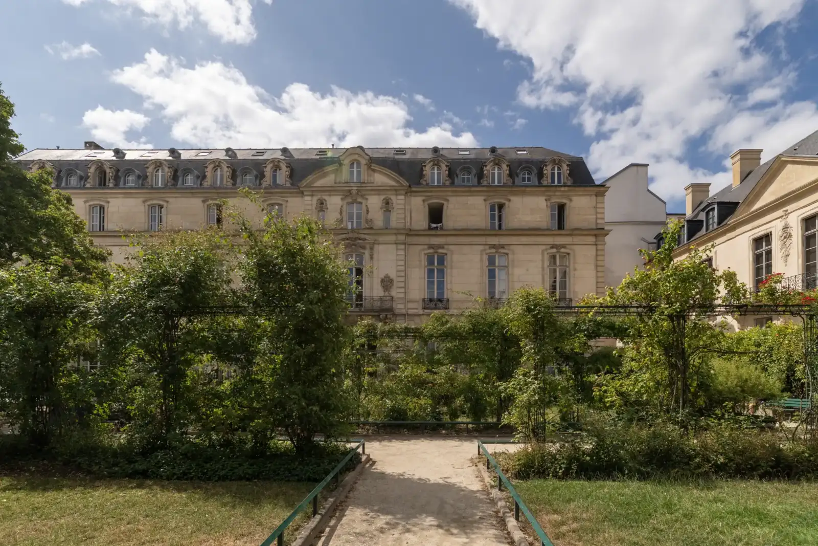 Vue depuis un jardin parisien arboré sur une élégante façade ancienne sous un ciel nuageux.