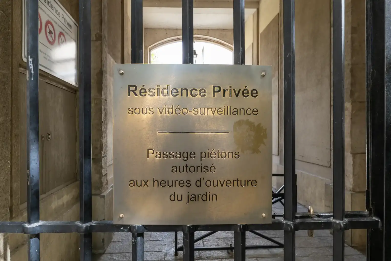 Une plaque signalétique métallique fixée sur une grille noire devant un passage voûté.