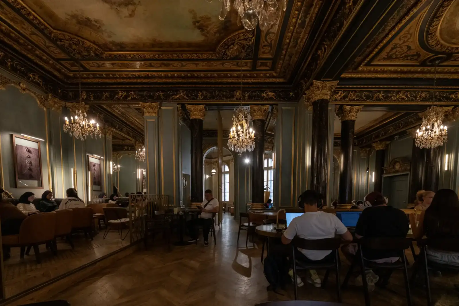 Des personnes sont attablées sous les plafonds dorés et somptueux d’un vaste café parisien.