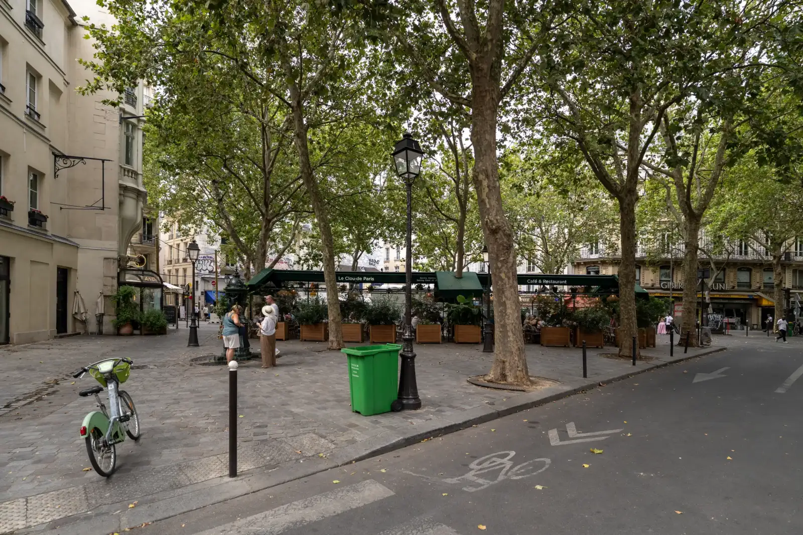 Une place parisienne ombragée avec des arbres, un café et des piétons.
