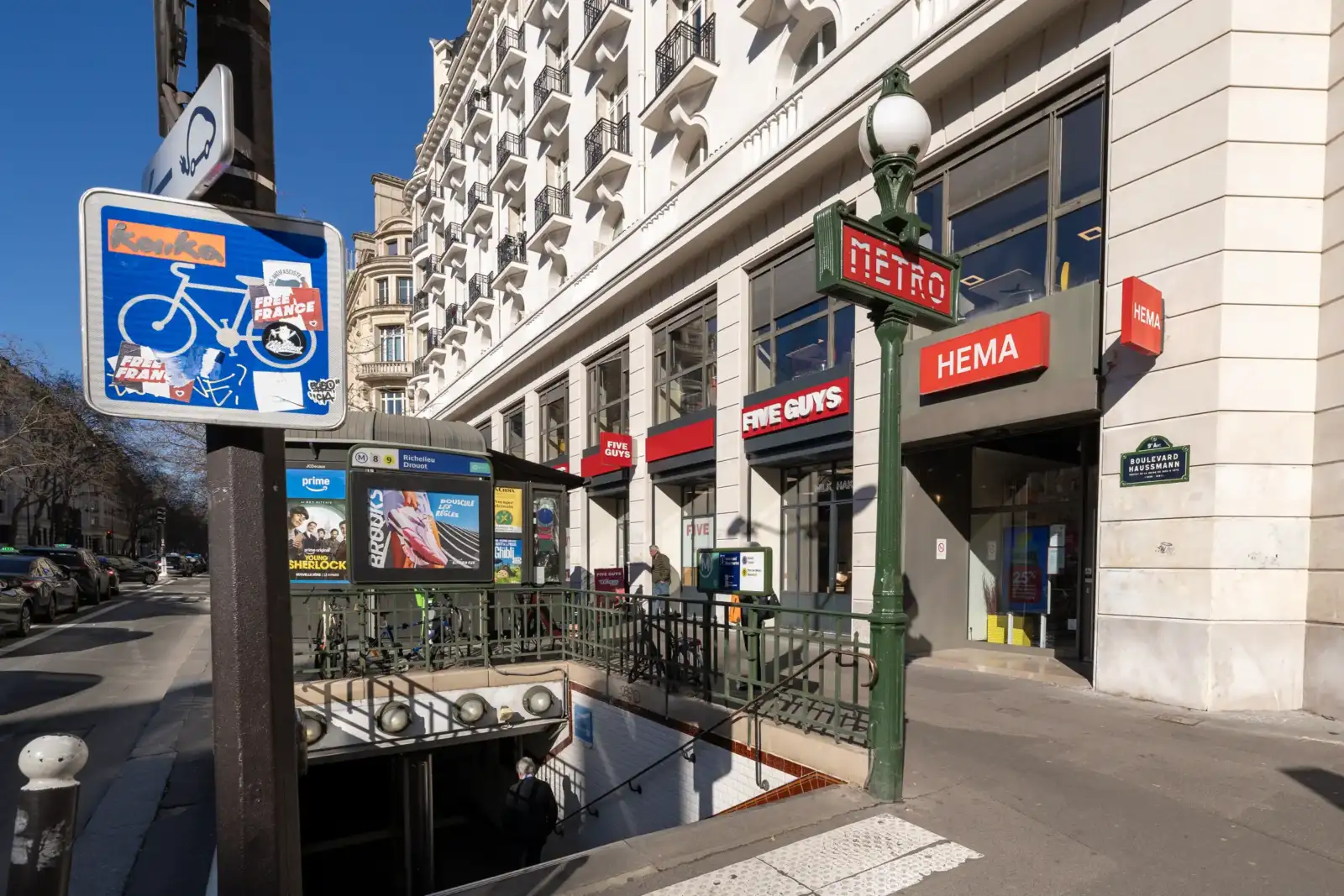 Une entrée de métro parisien avec sa signalétique classique devant des commerces haussmanniens ensoleillés.