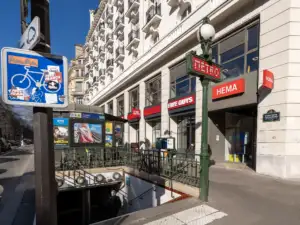 Une entrée de métro parisien avec sa signalétique classique devant des commerces haussmanniens ensoleillés.