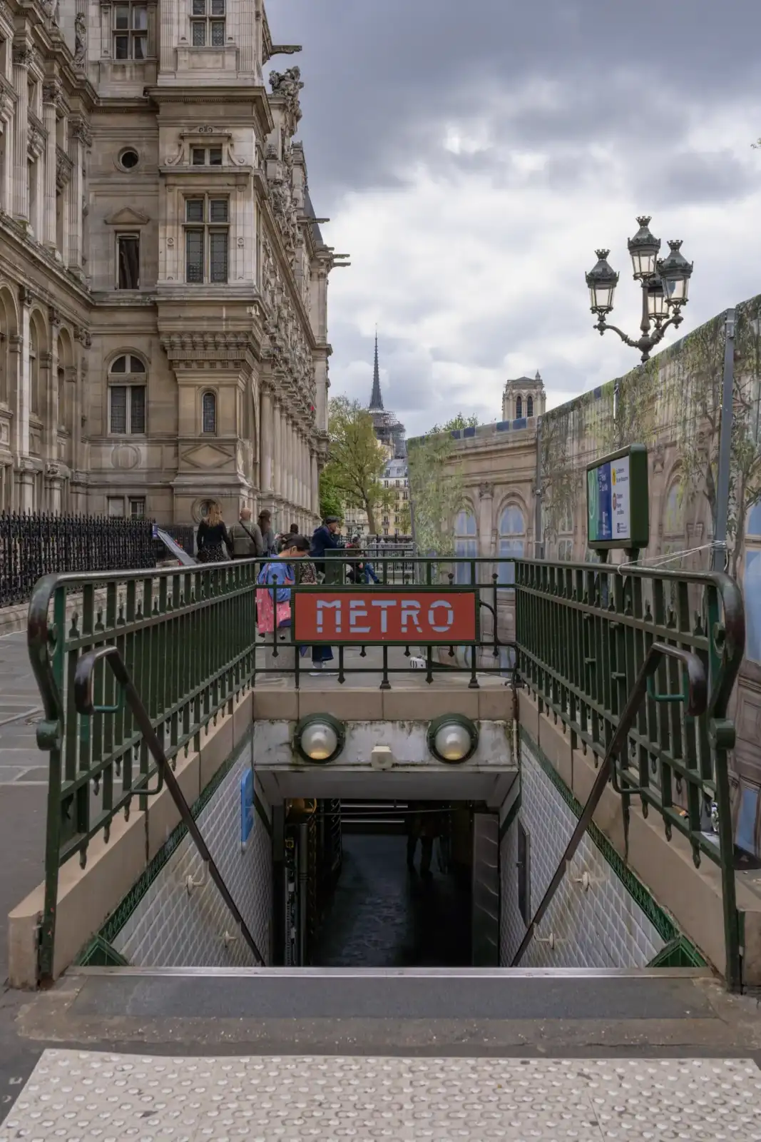 Une entrée de métro parisien avec l'Hôtel de Ville en arrière-plan sous un ciel gris.
