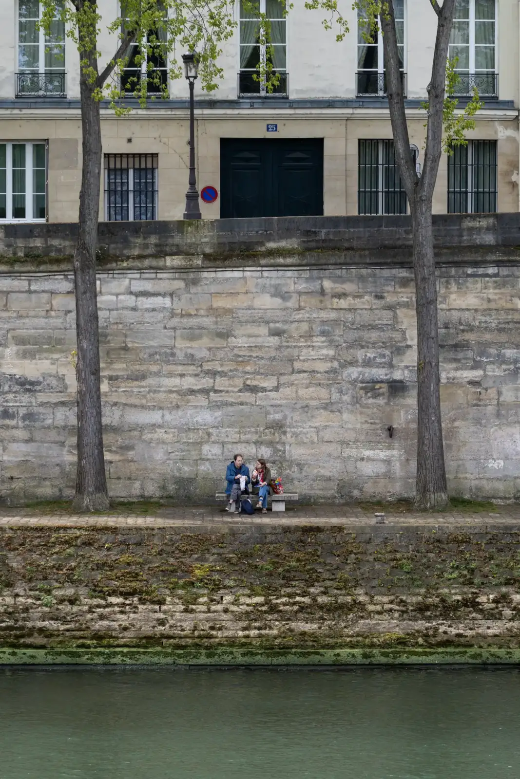 Un couple est assis sur un banc devant un haut mur en pierre parisien.