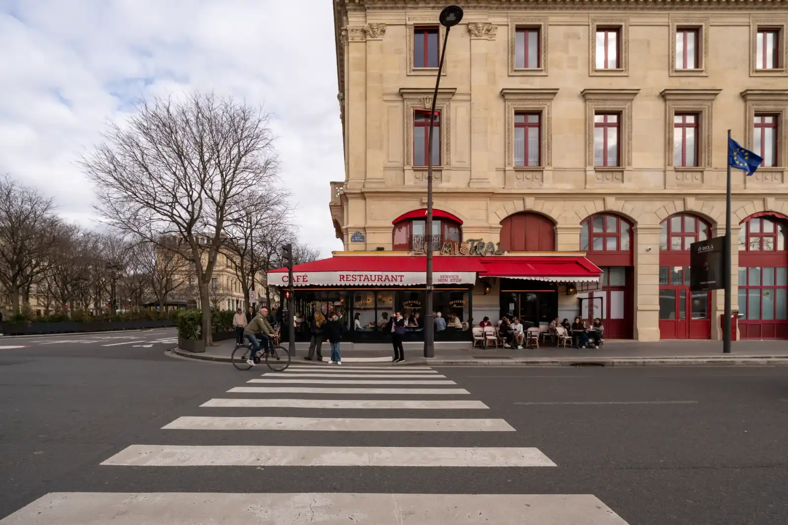 Une terrasse de café parisien en angle avec des clients sous un auvent rouge.