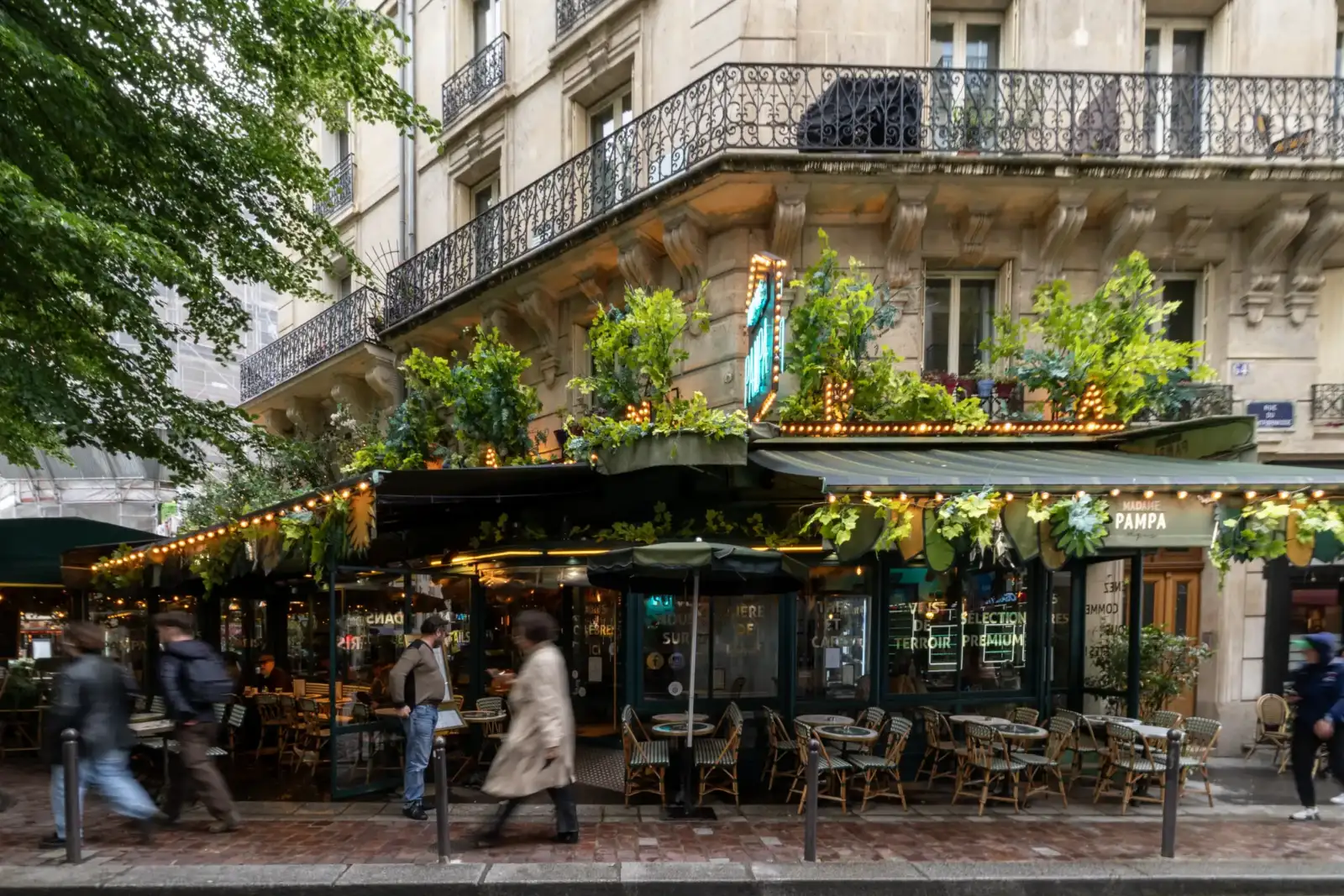 Une terrasse de café parisien végétalisée située au pied d'un immeuble en pierre.