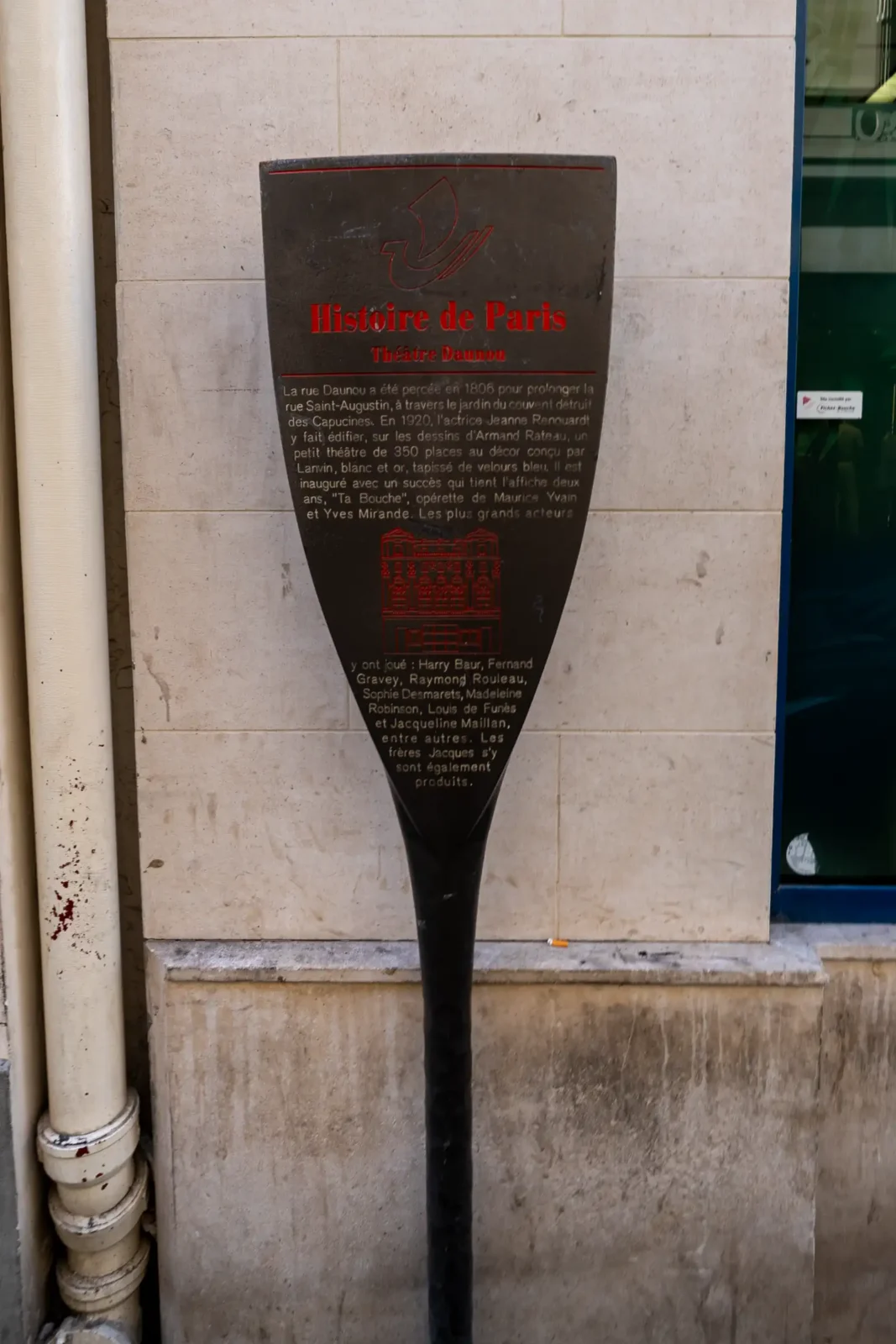Panneau informatif historique en forme de bouclier devant le Théâtre Daunou à Paris.