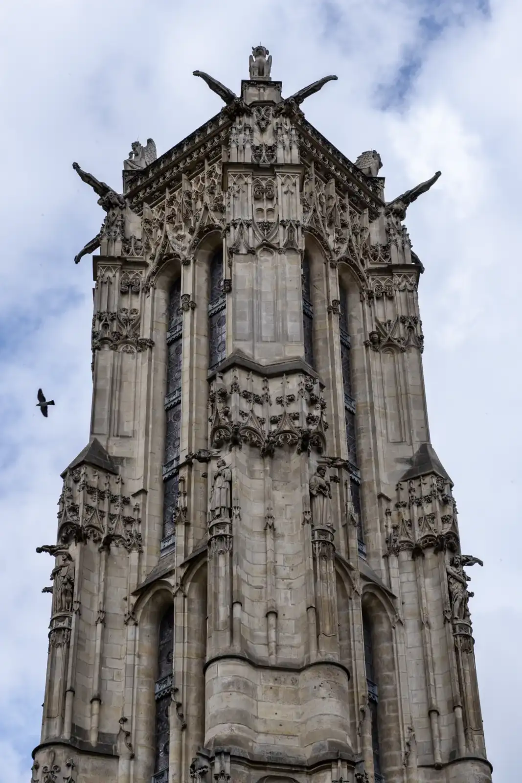La tour Saint-Jacques gothique s'élève vers un ciel nuageux avec un oiseau volant.
