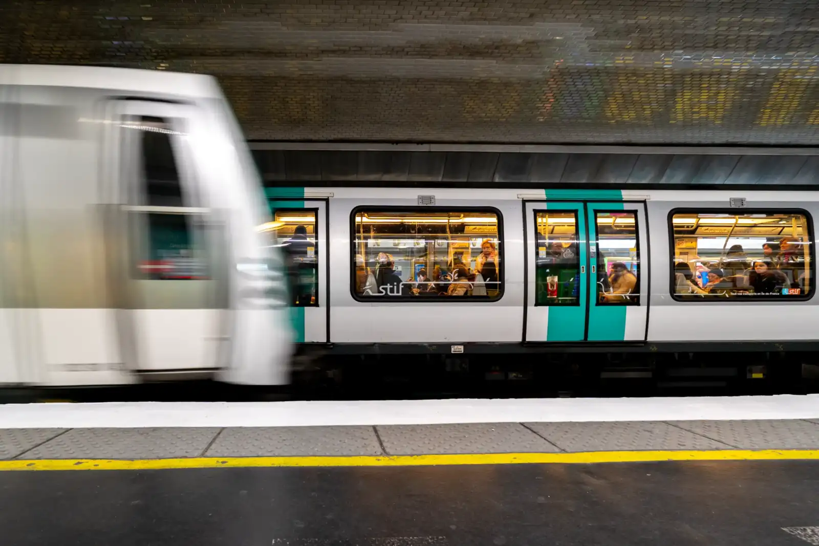 Un train de métro parisien passe rapidement devant un quai de station souterraine éclairée.