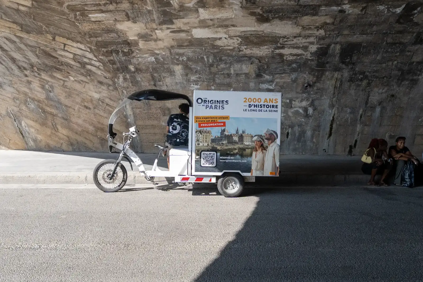 Un tricycle publicitaire pour des visites guidées historiques sous un pont parisien en pierre.