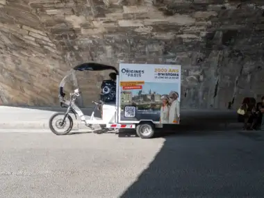 Un tricycle publicitaire pour des visites guidées historiques sous un pont parisien en pierre.