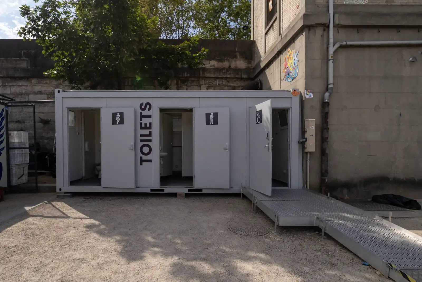 Une cabine de toilettes publiques en conteneur gris avec des portes ouvertes et une rampe d'accès.